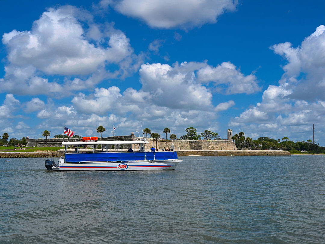A boat cruising along the water near a historic fort, with palm trees and a bright blue sky in the background.