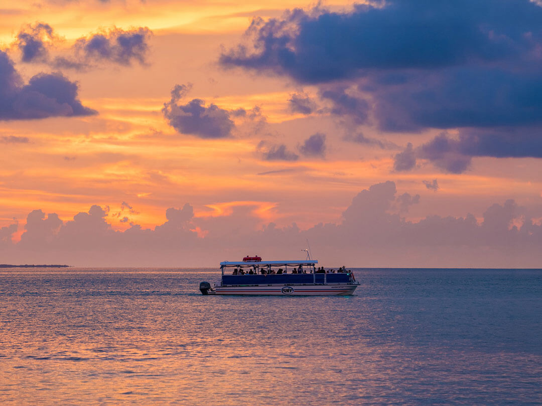A tour boat filled with passengers glides across calm water beneath a vivid orange and purple sunset sky streaked with dramatic clouds.