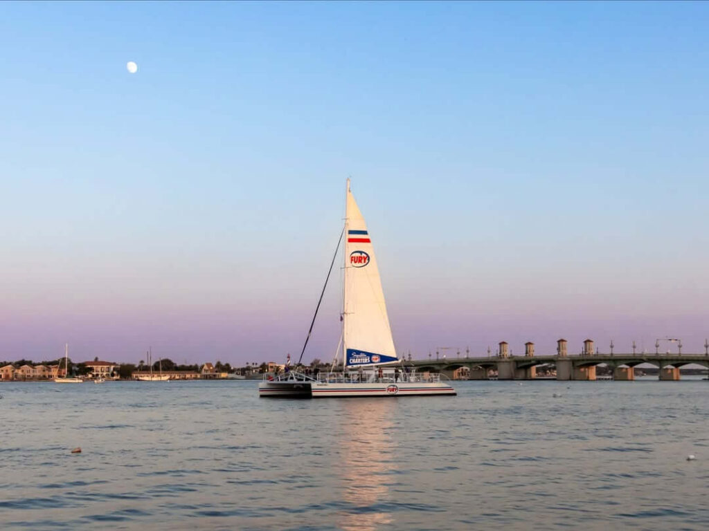 A sailing catamaran with its mainsail raised glides across calm water near the Bridge of Lions in St. Augustine, Florida, under a pastel pink and blue dusk sky with the moon visible above.
