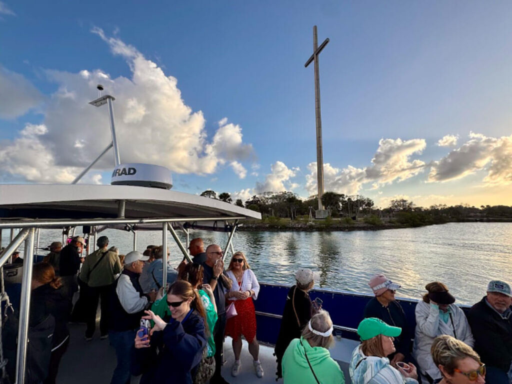 Passengers on a tour boat photograph a tall cross monument across the water at sunset in St. Augustine.