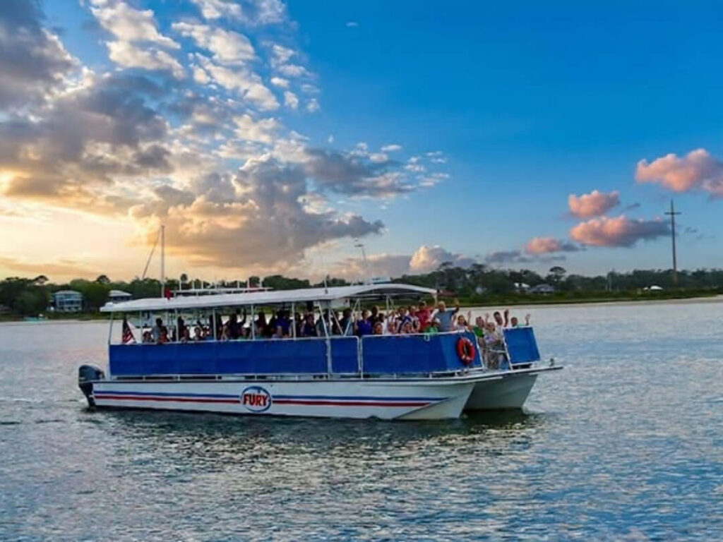 A double-hulled tour boat named Fury cruises the water packed with waving passengers under a colorful sunset sky.