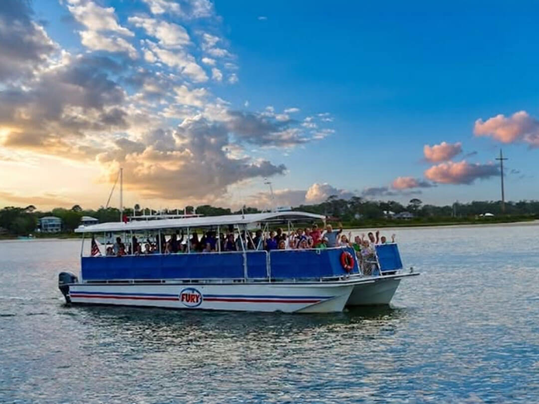 A double-hulled tour boat named Fury cruises the water packed with waving passengers under a colorful sunset sky.
