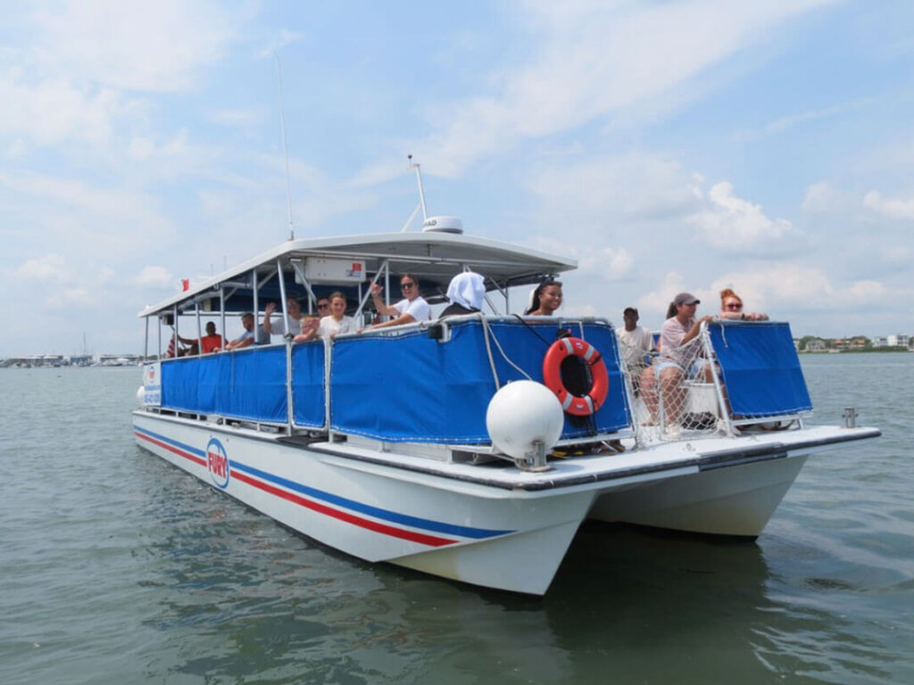 A white catamaran tour boat carrying a group of smiling, waving passengers cruises across calm waters under a daytime, cloudy blue sky.