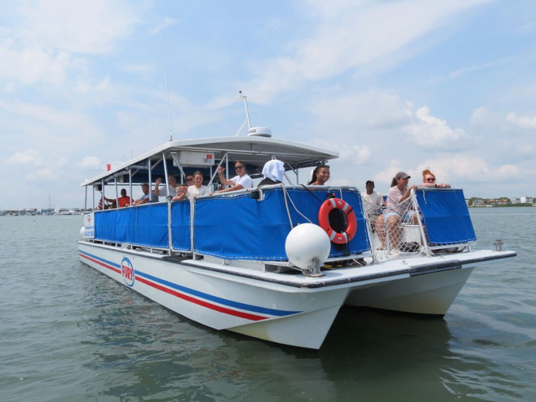 A white catamaran tour boat carrying a group of smiling, waving passengers cruises across calm waters under a daytime, cloudy blue sky.