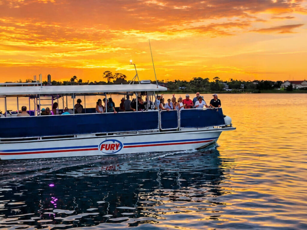 A white tour boat filled with passengers glides along the water against the backdrop of a vibrant orange and yellow sunset reflecting on the surface.
