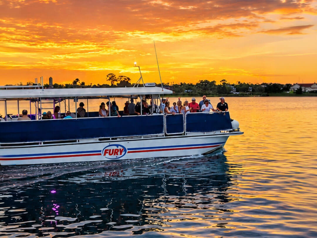 A white tour boat filled with passengers glides along the water against the backdrop of a vibrant orange and yellow sunset reflecting on the surface.