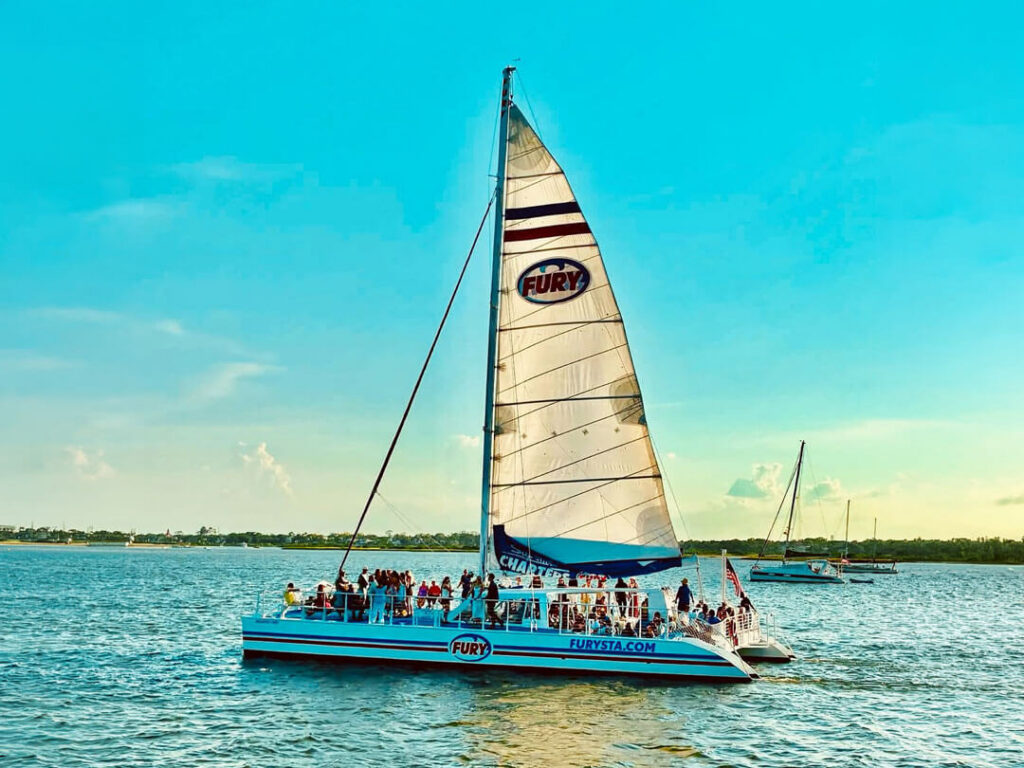 A sailboat catamaran full of passengers cruises the water under a large white sail.