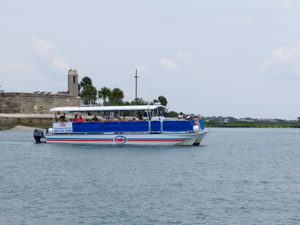 A tour boat carrying passengers passes a historic stone fort along the waterfront.